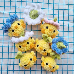 May include: A collection of handmade crochet chick ornaments in various styles. The chicks are primarily yellow with orange accents, black eyes, and some are adorned with blue, white, pink, and green floral or headwear details. They are arranged on a blue and white checkered surface.