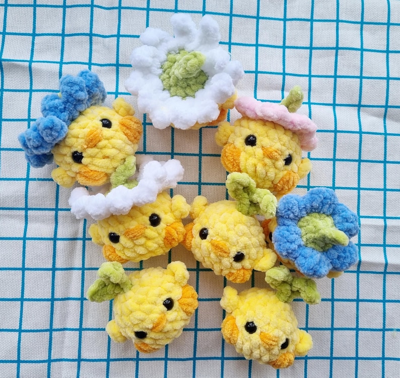 May include: A collection of handmade crochet chick ornaments in various styles. The chicks are primarily yellow with orange accents, black eyes, and some are adorned with blue, white, pink, and green floral or headwear details. They are arranged on a blue and white checkered surface.