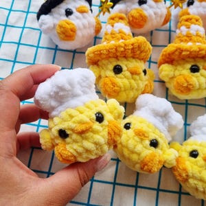 May include: A collection of small, handmade crochet plushies shaped like chicks. Some chicks wear white chef hats, while others wear orange patterned hats or black hats with gold stars. Each plush features bright yellow yarn and black eyes.