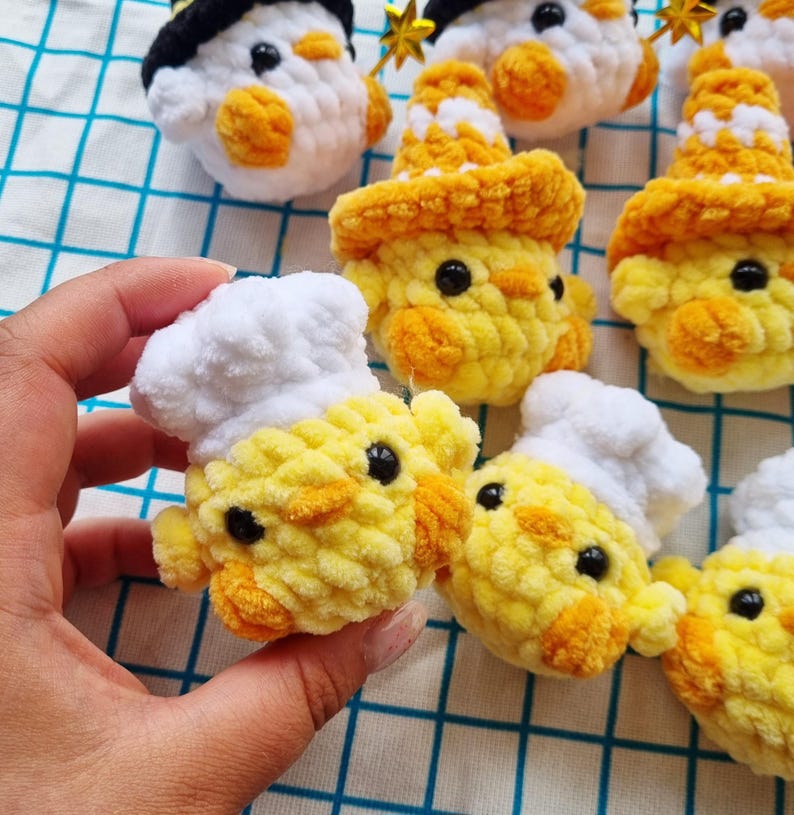 May include: A collection of small, handmade crochet plushies shaped like chicks. Some chicks wear white chef hats, while others wear orange patterned hats or black hats with gold stars. Each plush features bright yellow yarn and black eyes.