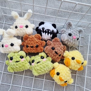 Puede incluir: Una colección de llaveros de animales de ganchillo hechos a mano, que incluyen conejos, un panda, un oso, una rana y una criatura amarilla. Cada llavero está hecho con hilo suave y texturizado y presenta ojos negros y detalles rosas.