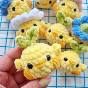 May include: A collection of handmade crochet chick ornaments in various colors. The chicks are primarily yellow with orange accents, black eyes, and some have green or blue floral details. The soft, textured yarn gives them a plush appearance.