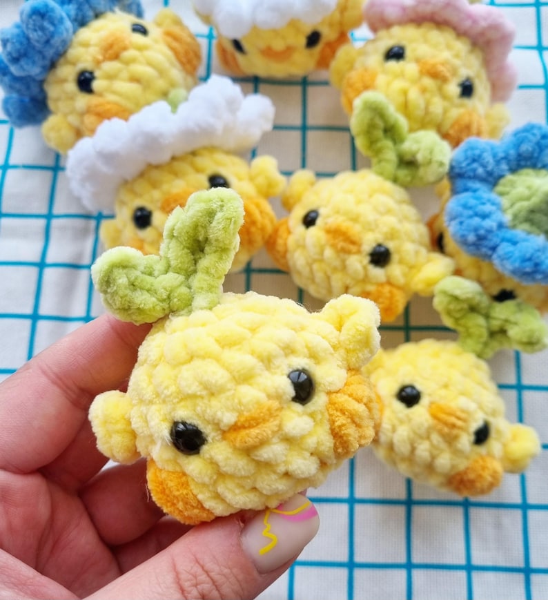 May include: A collection of handmade crochet chick ornaments in various colors. The chicks are primarily yellow with orange accents, black eyes, and some have green or blue floral details. The soft, textured yarn gives them a plush appearance.