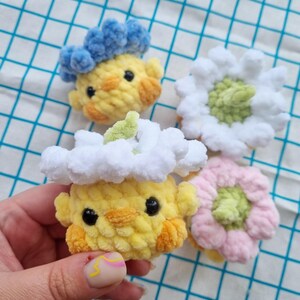 May include: Four handmade crochet items: two yellow chick-like figures with blue and white cloud-like hats, and two flower-shaped items in white and pink. The items have black eyes and orange accents. The background is a blue and white checkered pattern.