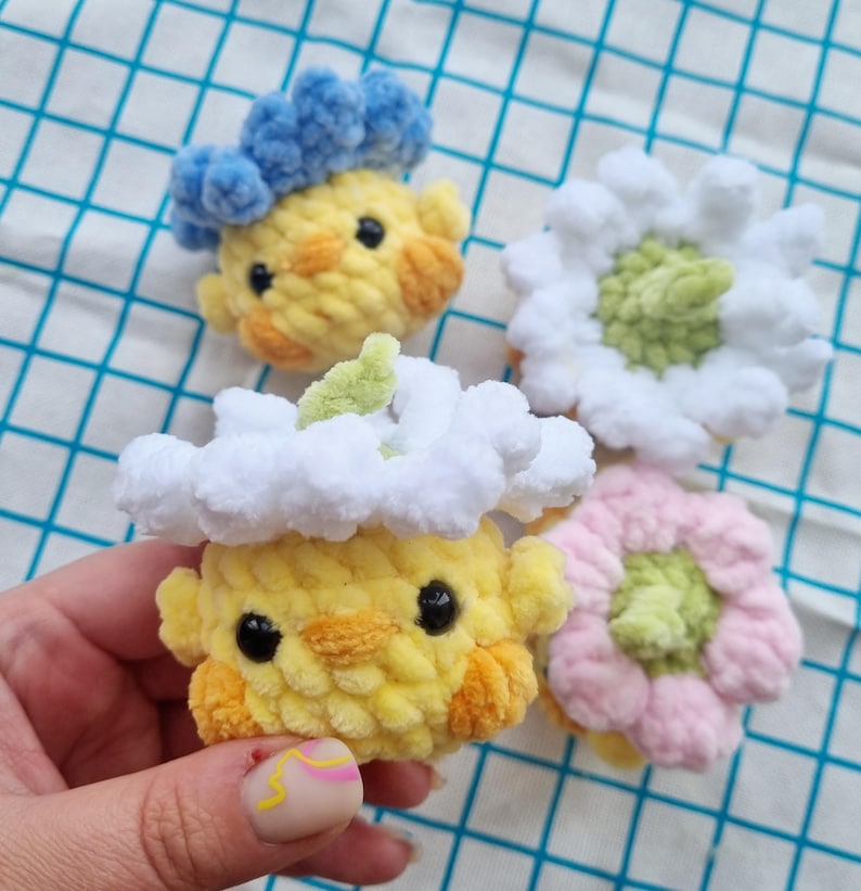 May include: Four handmade crochet items: two yellow chick-like figures with blue and white cloud-like hats, and two flower-shaped items in white and pink. The items have black eyes and orange accents. The background is a blue and white checkered pattern.