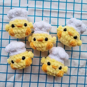 May include: Five small, round, yellow crocheted chicks, each wearing a white crocheted chef hat. The chicks have textured yarn bodies, orange beaks, and small black bead eyes. They are arranged in a group, showcasing their soft, plush appearance.