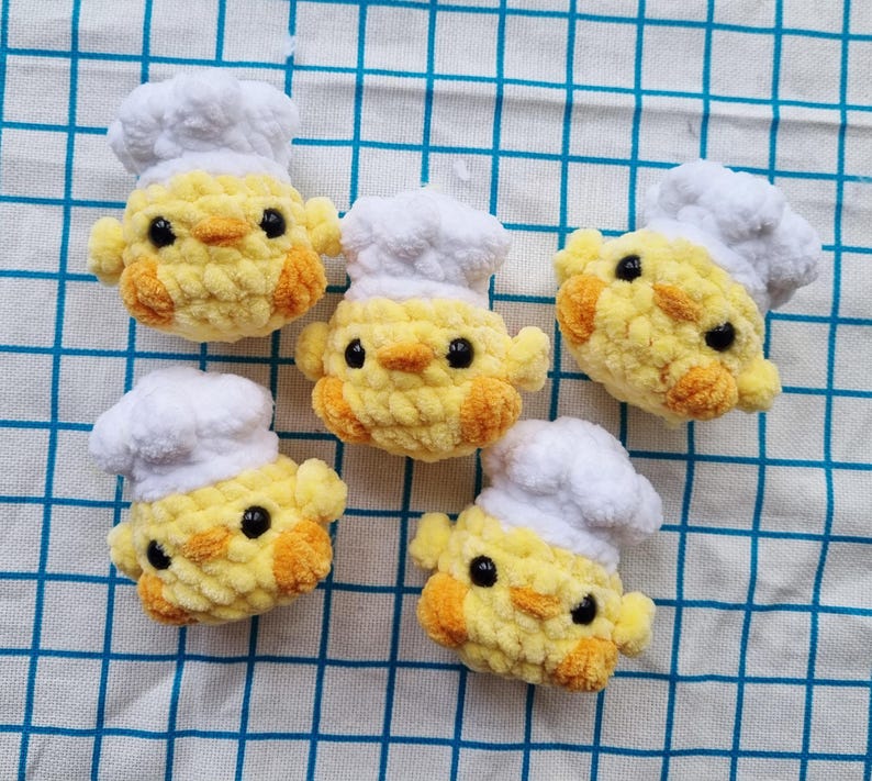 May include: Five small, round, yellow crocheted chicks, each wearing a white crocheted chef hat. The chicks have textured yarn bodies, orange beaks, and small black bead eyes. They are arranged in a group, showcasing their soft, plush appearance.