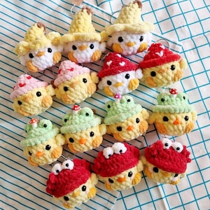 Op de afbeelding: Een verzameling van 15 gehaakte kuiken sleutelhangers met verschillende hoeden, waaronder een paddenstoel, een kikker, een hart en een banaan. De sleutelhangers zijn gemaakt van geel, groen, rood en wit garen.