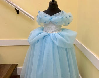 May include: A light blue princess dress on a black mannequin. The dress features a white bodice, a tulle skirt, and butterfly accents on the shoulders. A pearl-like belt encircles the waist. The dress is designed for a child.
