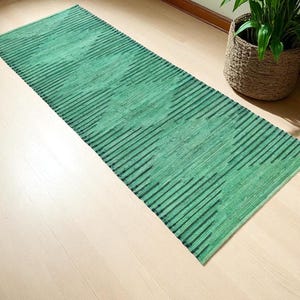 Puede incluir: Una alfombra rectangular verde con un patrón de rayas. La alfombra está colocada sobre un suelo de madera de color claro. Una cesta tejida con una planta verde está en el fondo.