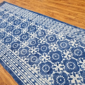 May include: A blue and white patterned rug with a floral design. The rug has a fringed edge and is made of cotton.