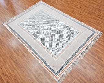 Puede incluir: Una alfombra de algodón blanco con un patrón geométrico negro y naranja. La alfombra tiene un borde de flecos y está hecha de fibras naturales.