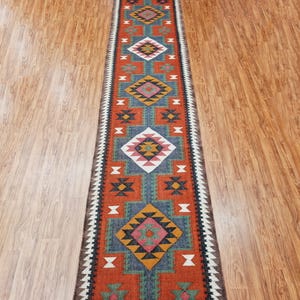 May include: A long, narrow rug with a geometric pattern in shades of red, orange, blue, green, and white. The rug has a fringe on both ends.