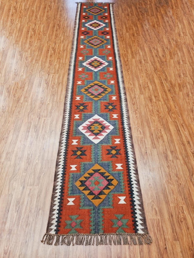 May include: A long, narrow rug with a geometric pattern in shades of red, orange, blue, green, and white. The rug has a fringe on both ends.