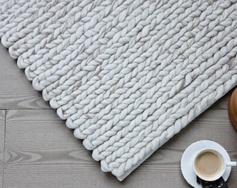 May include: A close-up of a chunky, off-white braided rug. The rug is made of thick, soft material and has a textured surface. A cup of coffee and a remote control are next to the rug.