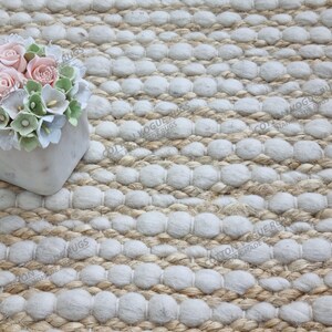 Könnte beinhalten: Nahaufnahme eines beigefarbenen und weißen strukturierten Teppichs mit einem Blumenarrangement. Der Teppich hat ein gewebtes Design mit Reihen weißer, kreisförmiger Büschel. Das Blumenarrangement befindet sich in einem quadratischen, hellfarbigen Topf und zeigt rosa und weiße Rosen.
