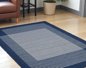 Op de afbeelding: Een rechthoekig marineblauw tapijt met een wit streepjespatroon. Het tapijt heeft een marineblauwe rand en een centraal gebied met dicht op elkaar geplaatste witte strepen. Het tapijt ligt op een houten vloer.