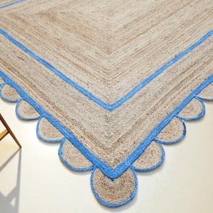 May include: A large, rectangular jute rug with a light brown color and a blue border. The border features a scalloped edge design. The rug is placed on a light-colored floor, and a small wooden table is visible in the corner.