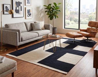 May include: A rectangular area rug featuring a bold geometric pattern of large black and cream-colored blocks. The rug is positioned on a wooden floor beneath a round wooden coffee table, a gray sofa, and a brown leather lounge chair with an ottoman.