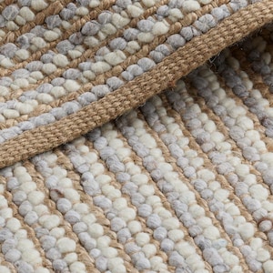 May include: Close-up of a woven rug with a textured surface. The rug has a striped pattern in shades of beige, white, and grey. The edges of the rug are finished with a thick, woven border.