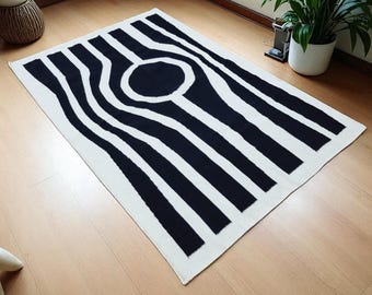 Puede incluir: Una alfombra rectangular con un patrón de alto contraste en blanco y negro. El diseño consiste en gruesas rayas verticales negras que se curvan hacia adentro para formar un círculo negro sólido en el centro de la alfombra, sobre un fondo blanco.