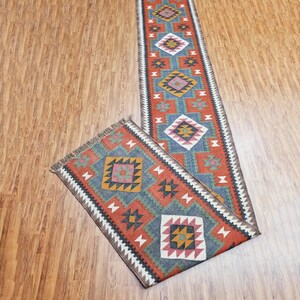 May include: A long, narrow rug with a geometric pattern in shades of red, blue, green, and brown. The rug has a fringe on both ends.