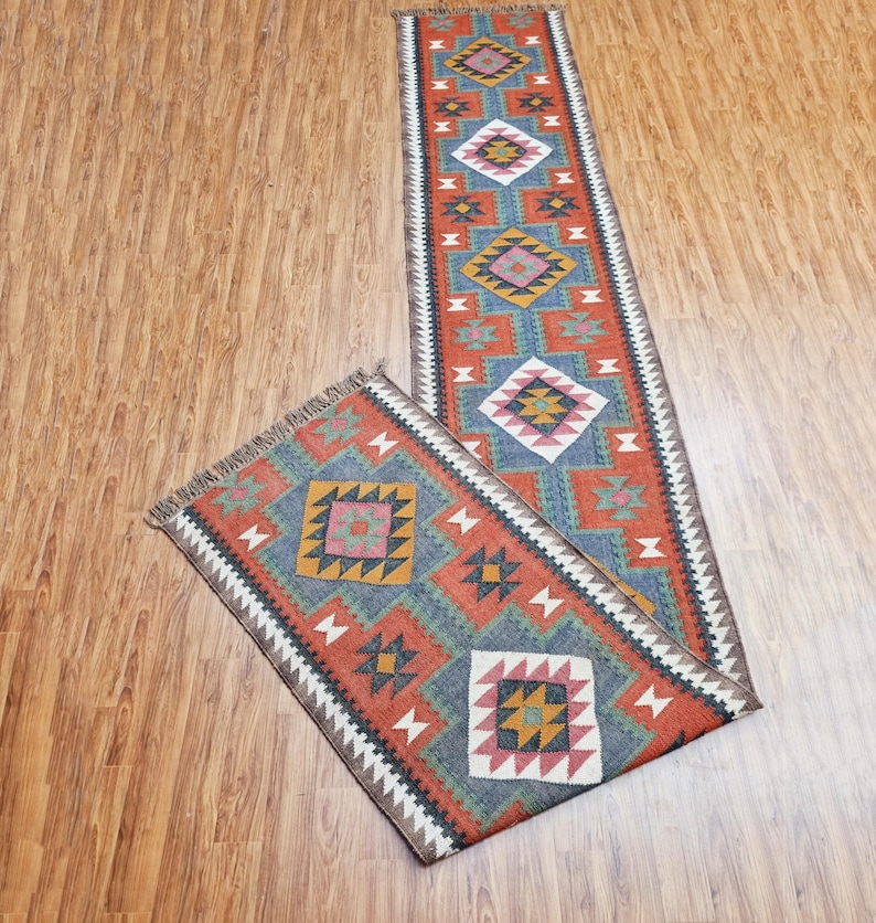 May include: A long, narrow rug with a geometric pattern in shades of red, blue, green, and brown. The rug has a fringe on both ends.
