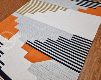 May include: A geometric patterned rug with black and white stripes, orange and beige accents. The rug has a white background and is made of cotton.