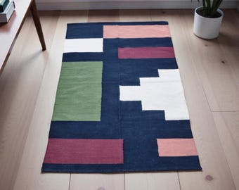 Puede incluir: Una alfombra rectangular con un diseño geométrico. La alfombra presenta un fondo azul oscuro con bloques blancos, verdes, rosas y burdeos. Está colocada sobre un suelo de madera clara.