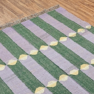 Könnte beinhalten: Ein handgewebter Teppich mit einem Streifenmuster in Grün und Lavendel. Der Teppich hat ein sich wiederholendes Design aus gelben Blattformen. Der Teppich hat einen Fransenrand und liegt auf einem Holzboden.