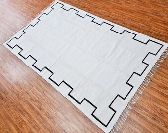 May include: White rug with a black geometric border design. The rug has fringe along the edges.