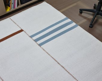 Op de afbeelding: Witte rechthoekige vloerkleed met bruine en blauwe strepen. Het vloerkleed heeft een eenvoudig ontwerp met een bruine streep diagonaal over de linkerbovenhoek en drie blauwe strepen aan de rechterkant. Het vloerkleed is gemaakt van geweven materiaal.