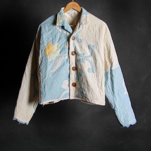 May include: A white and light blue quilted jacket with a floral pattern. The jacket has a button-up front and long sleeves.