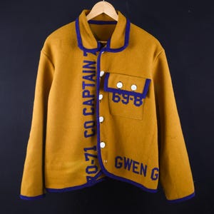 May include: Mustard yellow jacket with blue trim and lettering. The jacket features the text "70-71 CO CAPTAIN 7" vertically down the front, and "69-8" and "GWEN G" on the pocket. It has white buttons and a collar.