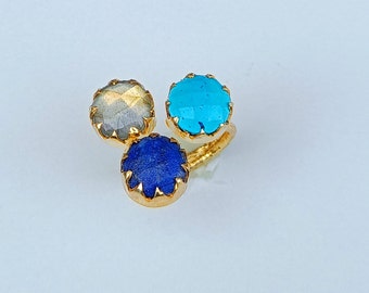 May include: A gold ring featuring three gemstone settings. The ring has a turquoise, a blue, and a gray gemstone. The gemstones are round and set in a gold bezel. The ring is on a white background.