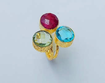Könnte beinhalten: Ein goldfarbener Ring mit drei Edelsteinen: einem Rubin, einem hellgrünen Stein und einem blauen Stein. Die Edelsteine sind in strukturierten Goldfassungen gefasst. Der Ring hat ein einzigartiges Drei-Stein-Design.
