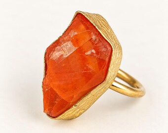 May include: A gold-toned ring featuring a large, irregular, faceted orange stone set within a textured metal bezel. The band is smooth and circular, contrasting with the rough, organic appearance of the setting that holds the vibrant, translucent orange gemstone securely in place.