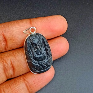 Może przedstawiać: Czarny, owalny wisiorek ze srebrnym uchwytem. Wisiorek przedstawia rzeźbiony wzór postaci. Wisiorek jest trzymany w dłoni osoby na ciemnym tle. Wisiorek jest prawdopodobnie wykonany z kamienia.