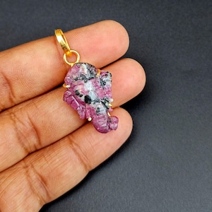 May include: A gold-colored pendant with a carved ruby elephant design. The ruby is a deep pink color with black and white inclusions. The pendant is suspended from a gold-colored bail.