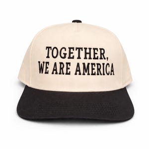 May include: A baseball cap with a cream-colored crown and a black brim. The cap features the embroidered text "TOGETHER, WE ARE AMERICA" in bold, black letters. The cap has a black button on top.