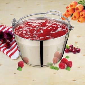 May include: A stainless steel jam pan with a handle, filled with red jam. The pan is sitting on a wooden surface with raspberries and cherries scattered around it.