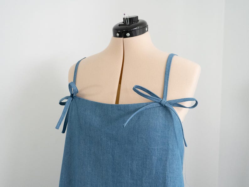 K&ouml;nnte beinhalten: Ein blaues, &auml;rmelloses Kleid aus Denim mit verstellbaren Tr&auml;gern, die an den Schultern zu Schleifen gebunden werden.