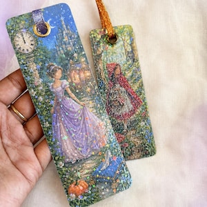 May include: Two rectangular bookmarks featuring fairy tale illustrations with a holographic star finish. One depicts a figure in a purple gown with a clock, pumpkin, and carriage. The other shows a figure in a red hooded cloak in a forest.