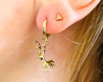 May include: A close-up of a person's ear adorned with gold earrings. One earring is a small gold triangle stud. The other is a gold hoop with a detailed deer pendant. The jewelry is set against the backdrop of the person's skin and hair.