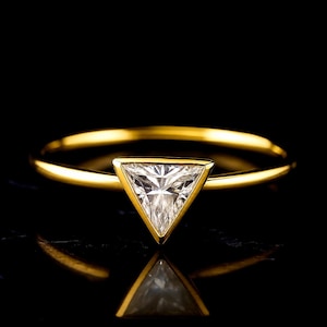 Peut inclure: Une bague en or ornée d'une pierre précieuse transparente taillée en triangle, fixée dans un serti clos. L'anneau est fin et poli, présentant une surface métallique lisse et réfléchissante qui contraste avec la pierre blanche facettée.