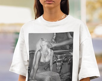 May include: White t-shirt featuring a black and white image of a woman in a dress. The woman is sitting in a chair, and a hand is touching her face. The t-shirt is worn by a person wearing black sunglasses.