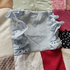 Puede incluir: Una blusa azul claro con mangas con volantes y detalles de ojal. La blusa est&aacute; sobre un fondo de patchwork de varias telas en tonos beige, verde, rosa, rojo y blanco.