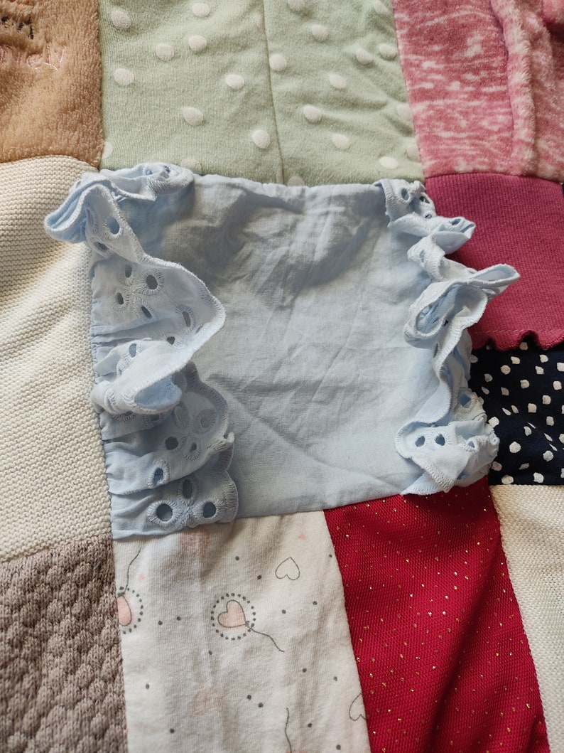 Puede incluir: Una blusa azul claro con mangas con volantes y detalles de ojal. La blusa est&aacute; sobre un fondo de patchwork de varias telas en tonos beige, verde, rosa, rojo y blanco.
