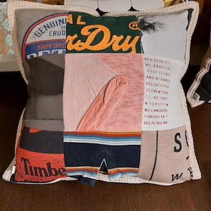 Peut inclure: Coussin carré en patchwork fait à partir de t-shirts recyclés. Le coussin présente diverses couleurs et graphismes, notamment des textes des marques "Superdry" et "Timberland". Le coussin a une bordure crème et est posé sur une surface en bois.