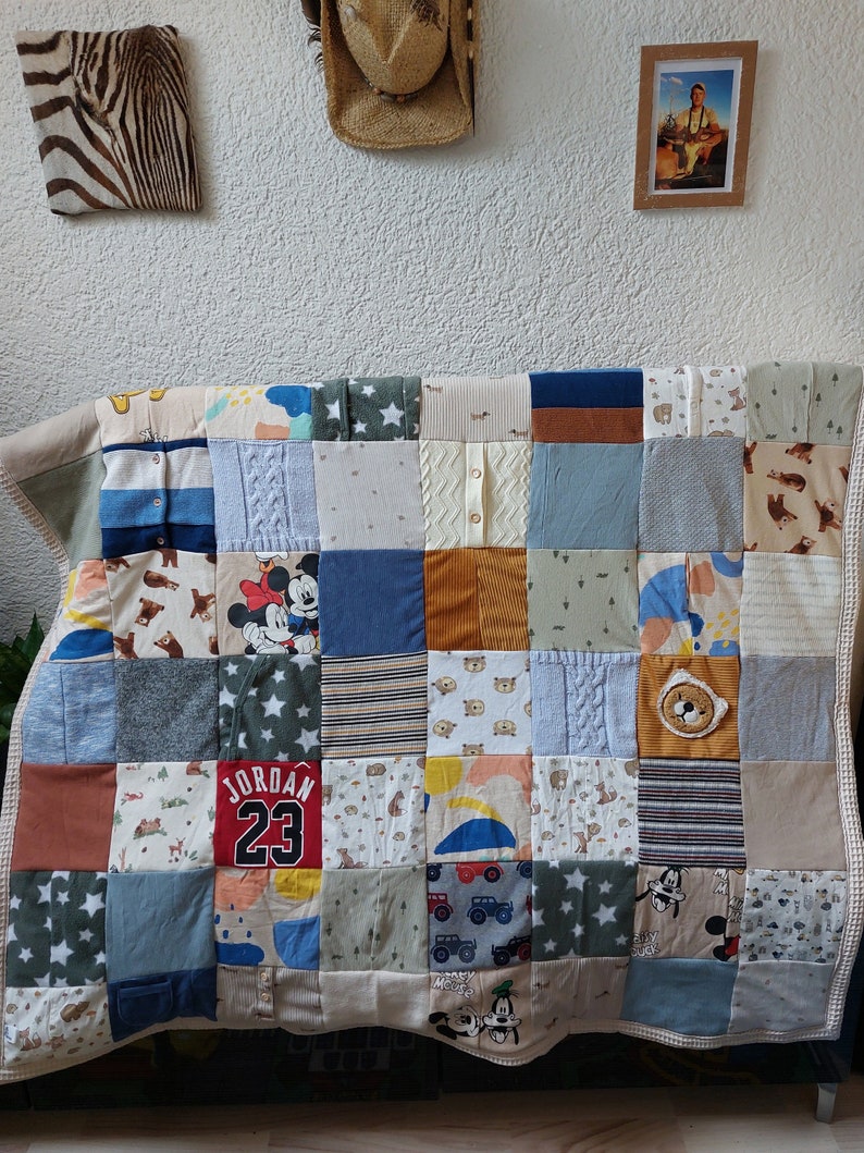 Puede incluir: Un edred&oacute;n de retazos hecho con cuadrados de tela variados. Los cuadrados presentan diferentes patrones, incluyendo estrellas, osos y Mickey Mouse. Algunos cuadrados est&aacute;n hechos de ropa, incluyendo una camiseta con "JORDAN 23". El edred&oacute;n est&aacute; sobre un sof&aacute;.